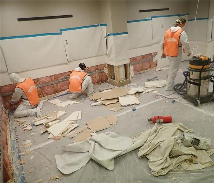 four workers with protective gear removing drywall from a building, flood cuts performed, debris on the floor.